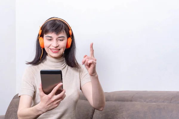 Mujer atractiva sentada en un sofá con auriculares naranjas mirando dentro de una tableta y levantando el dedo — Foto de Stock