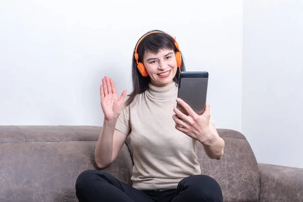 Encantadora morena con auriculares naranjas haciendo una videollamada y mostrando un gesto de "hola o adiós". El uso de dispositivos electrónicos portátiles para el chat de vídeo distante, llamando a los padres. Nuevo sin concepto — Foto de Stock