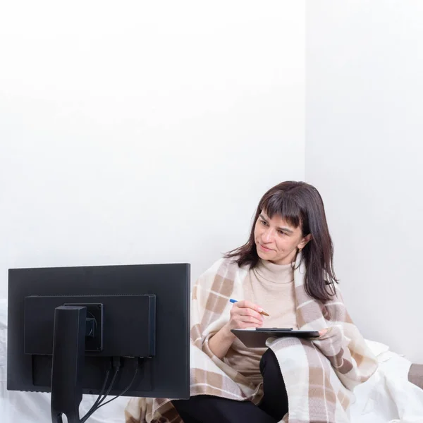 Una mujer atractiva cubierta con una manta sosteniendo un cuaderno y un bolígrafo y mirando de cerca a un monitor — Foto de Stock
