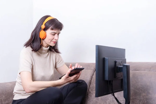 Una mujer sentada en un sofá con auriculares naranjas frente a un monitor de ordenador y presionando una tableta. — Foto de Stock