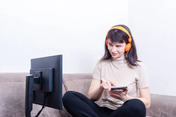 Mujer atractiva con auriculares naranjas mirando un monitor de computadora y presionando una tableta con su dedo — Foto de Stock