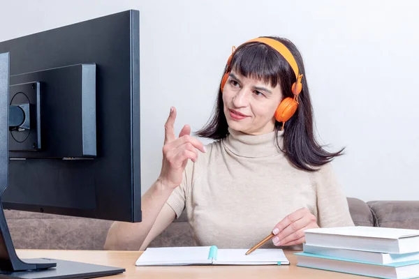 Una mujer con auriculares naranjas sentada en una mesa con libros, mirando un monitor de computadora, levantando el dedo — Foto de Stock