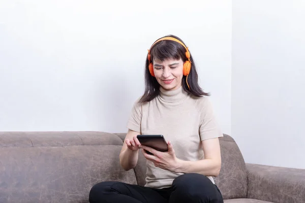 Mujer atractiva sentada en un sofá con auriculares naranjas escribiendo texto en una tableta — Foto de Stock