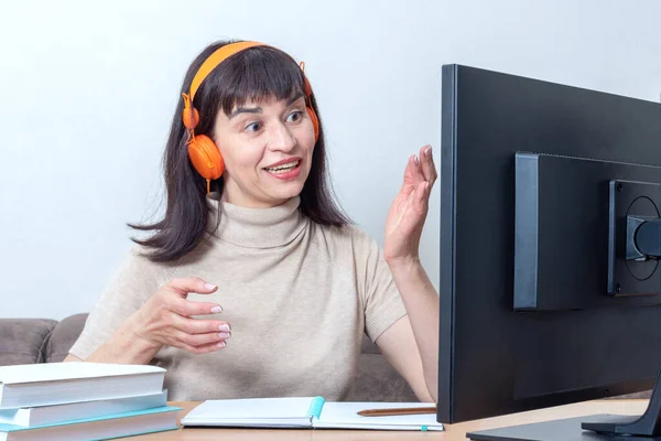 Una atractiva profesora de auriculares naranjas sentada frente a un monitor de ordenador, explica una tarea en línea — Foto de Stock
