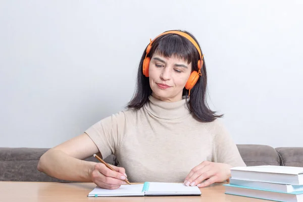Estudiante sonriente escuchando un curso de audio remoto en auriculares y escribiendo algo en un cuaderno mientras está sentada a la mesa. Enseñanza a distancia. Aprendizaje electrónico. — Foto de Stock