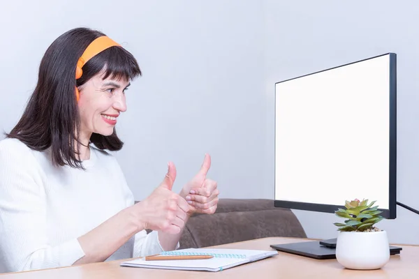 Mujer de negocios sonriente con auriculares mira en un monitor con una pantalla blanca en blanco y sostiene pulgares hacia arriba — Foto de Stock