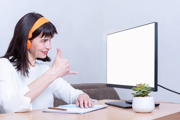 Mujer de negocios sonriente en auriculares mira en un monitor con una pantalla blanca en blanco y muestra un signo de llamada — Foto de Stock