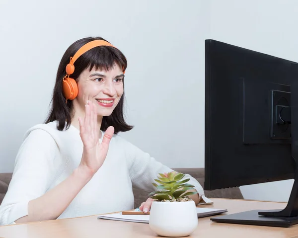 Mujer encantadora con auriculares naranjas con una videollamada y un gesto de bienvenida frente a un monitor de computadora. — Foto de Stock