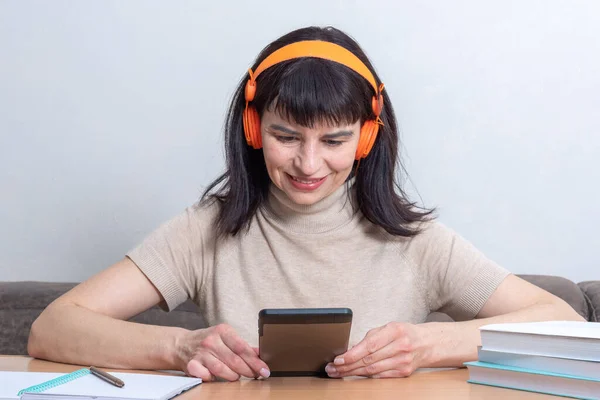 Mujer feliz con auriculares sonríe y navega por las redes sociales en la tableta, sentada a la mesa con libros y escuchando música o audiolibro en casa. Enseñanza a distancia. Aprendizaje electrónico. — Foto de Stock