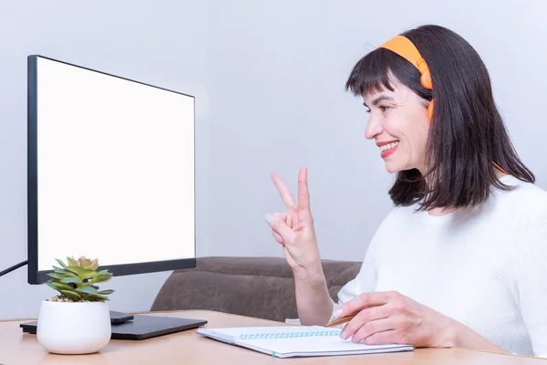 Mujer de negocios sonriente con auriculares mira en un monitor con una pantalla blanca en blanco y muestra una señal de victoria — Foto de Stock
