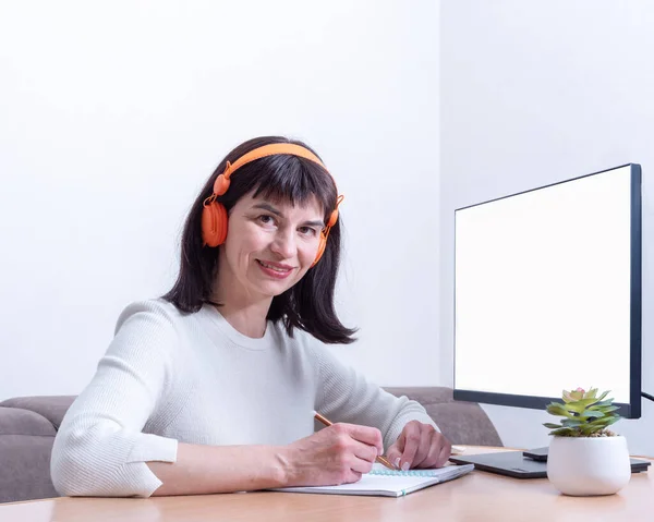 Mujer de negocios sonriente con auriculares te mira mientras estás sentado en la mesa frente a un monitor con una pantalla en blanco — Foto de Stock