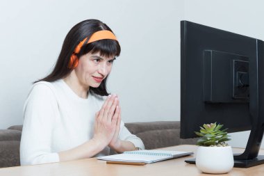 Beyaz kazaklı ve kulaklıklı bir kadın monitörün önündeki masada oturuyor ve bir teşekkür işareti gösteriyor.