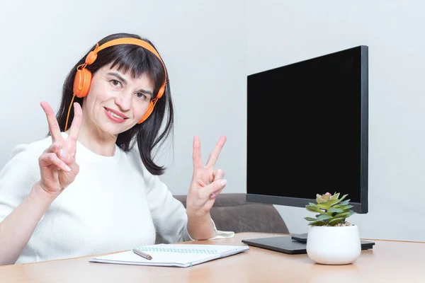 Mujer de negocios sonriente con auriculares te mira, sentada en la mesa frente al monitor con una pantalla negra — Foto de Stock