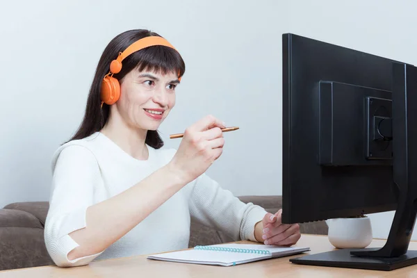 Smiling woman in headphones sitting at the table looking at the monitor screen, talking on video communication