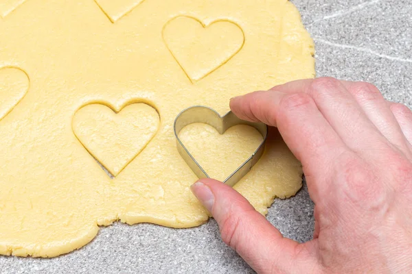 Banketbakkers hand snijdt hartvormige koekjes van rauw geel deeg op een grijze tafelachtergrond, close-up. — Stockfoto