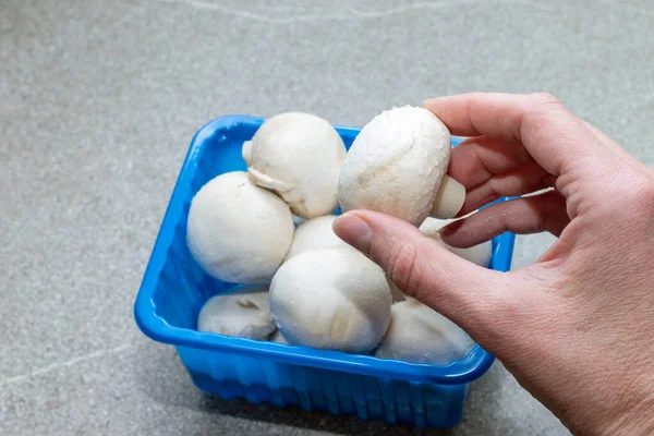 Una mano saca un Champignon Mushroom de una caja azul en una mesa de cocina gris, de cerca. Champiñones blancos enteros en una caja de plástico. Comida natural saludable. Concepto de alimentación dietética — Foto de Stock