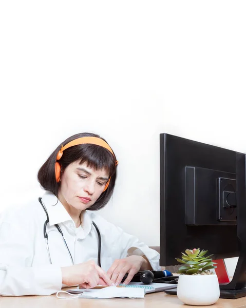 A female doctor wears a headset makes an online video call with a patient, signs a document in her office, copy space