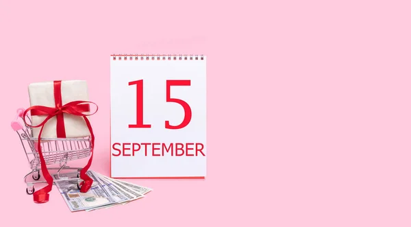 Pudełko prezentów w wózku sklepowym, dolarach i kalendarzu z datą 15 września na różowym tle. — Zdjęcie stockowe