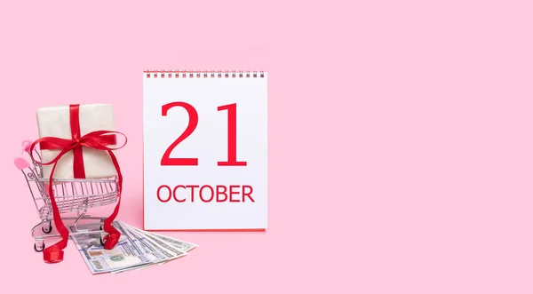 Eine Geschenkbox im Einkaufswagen, Dollars und ein Kalender mit dem Datum 21. Oktober auf rosa Hintergrund. — Stockfoto