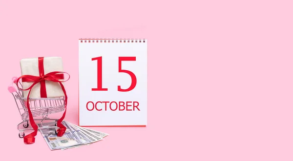 Eine Geschenkbox im Einkaufswagen, Dollars und ein Kalender mit dem Datum des 15. Oktober auf rosa Hintergrund. — Stockfoto
