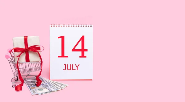 En presentask i en kundvagn, dollar och en kalender med datum 14 juli på en rosa bakgrund. — Stockfoto