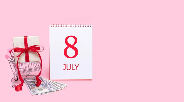 En presentask i en kundvagn, dollar och en kalender med datum 8 juli på en rosa bakgrund. — Stockfoto