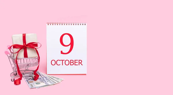 Pudełko prezentów w wózku sklepowym, dolarach i kalendarzu z datą 9 października na różowym tle. — Zdjęcie stockowe