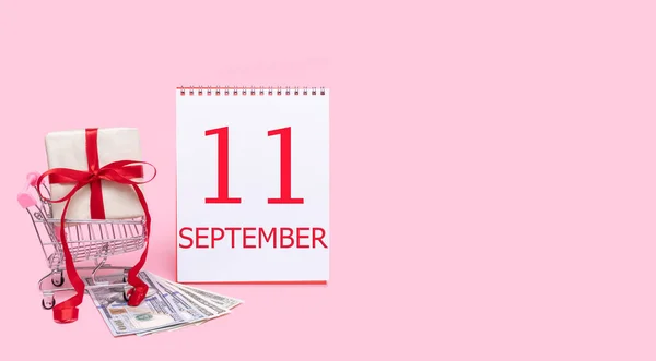 Eine Geschenkbox im Einkaufswagen, Dollars und ein Kalender mit dem Datum des 11. September auf rosa Hintergrund. — Stockfoto
