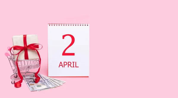 Una caja de regalo en un carrito de la compra, dólares y un calendario con la fecha del 2 de abril sobre un fondo rosa. — Foto de Stock