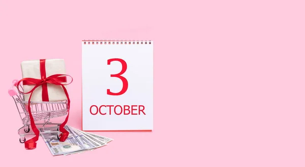 Eine Geschenkbox im Einkaufswagen, Dollars und ein Kalender mit dem Datum 3. Oktober auf rosa Hintergrund. — Stockfoto