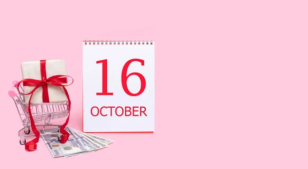 Ένα κουτί δώρου σε ένα καροτσάκι με ψώνια, δολάρια και ένα ημερολόγιο με ημερομηνία 16 Οκτωβρίου σε ροζ φόντο.. — Φωτογραφία Αρχείου