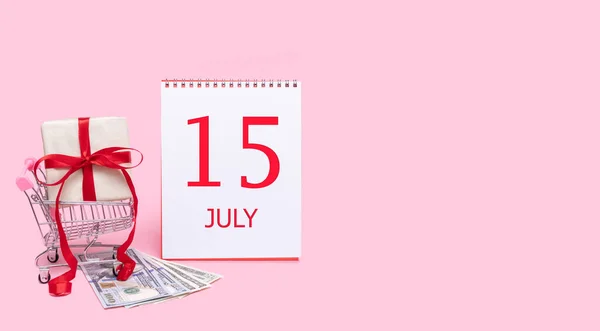 En presentask i en kundvagn, dollar och en kalender med 15 juli på rosa bakgrund. — Stockfoto