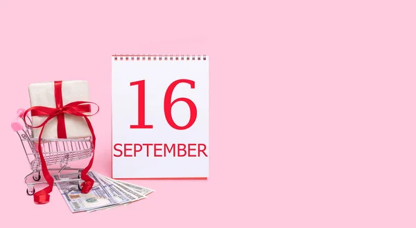 Pudełko prezentów w wózku sklepowym, dolarach i kalendarzu z datą 16 września na różowym tle. — Zdjęcie stockowe