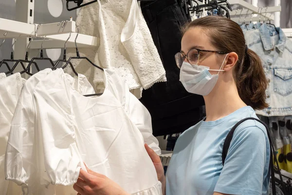 Una hermosa joven con una máscara médica para protegerse del coronavirus está eligiendo ropa en una tienda — Foto de Stock