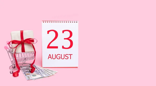 En presentask i en kundvagn, dollar och en kalender med datum 23 augusti på en rosa bakgrund. — Stockfoto