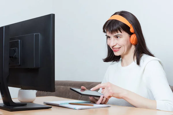 Una mujer sonriente con auriculares naranjas sentada frente al monitor y sosteniendo un smartphone para ir de compras — Foto de Stock
