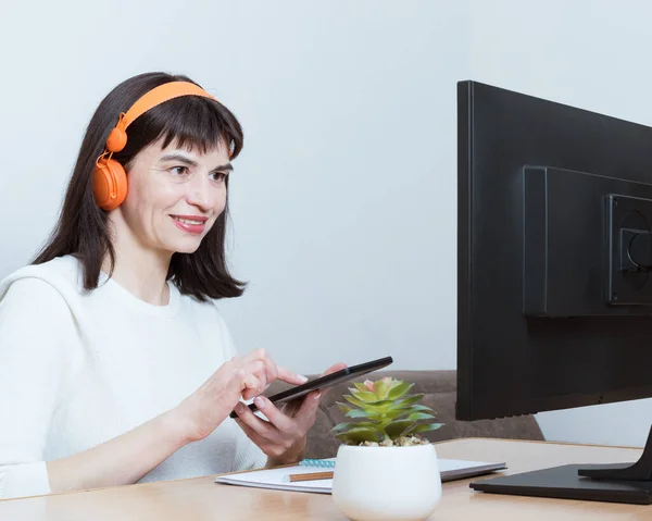 Una mujer con auriculares sentada frente al monitor y sosteniendo un smartphone para la búsqueda de información — Foto de Stock