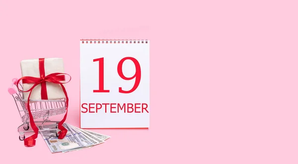 Pudełko prezentów w wózku sklepowym, dolarach i kalendarzu z datą 19 września na różowym tle. — Zdjęcie stockowe