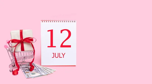 En presentask i en kundvagn, dollar och en kalender med datumet 12 juli på en rosa bakgrund. — Stockfoto