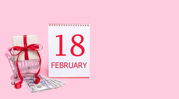 Una caja de regalo en un carrito de la compra, dólares y un calendario con la fecha del 18 de febrero sobre un fondo rosa. —  Fotos de Stock