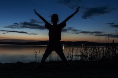 Gün batımında gölün kıyısında kollarını kaldırıp zıplayan bir çocuğun silueti. Göl kenarında aile tatili, kamp.