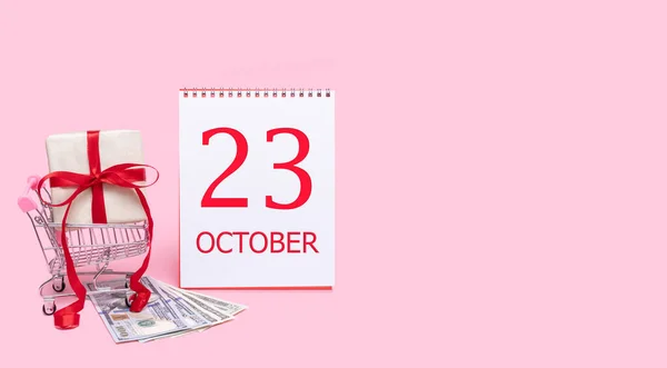 Pudełko prezentów w wózku sklepowym, dolarach i kalendarzu z datą 23 października na różowym tle. — Zdjęcie stockowe
