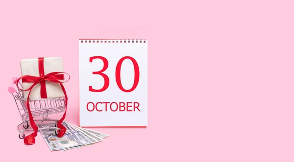 Eine Geschenkbox im Einkaufswagen, Dollars und ein Kalender mit dem Datum des 30. Oktober auf rosa Hintergrund. — Stockfoto