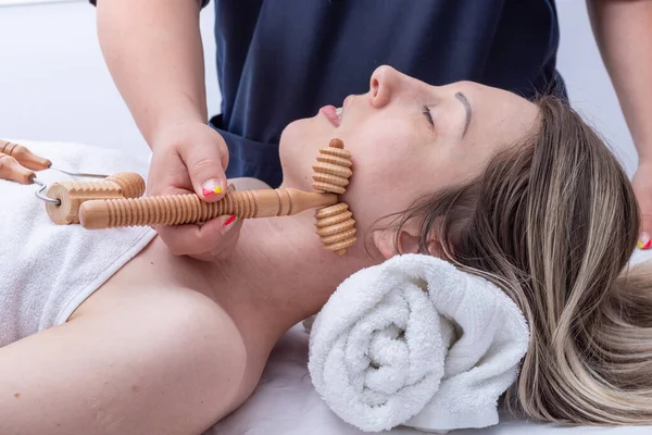 Madero terapia, masaje de cara y cuello, masaje relajante antienvejecimiento - Las manos del masajista masajea la cara y el cuello de la niña utilizando un masajeador de madera con pinchos, primer plano. —  Fotos de Stock