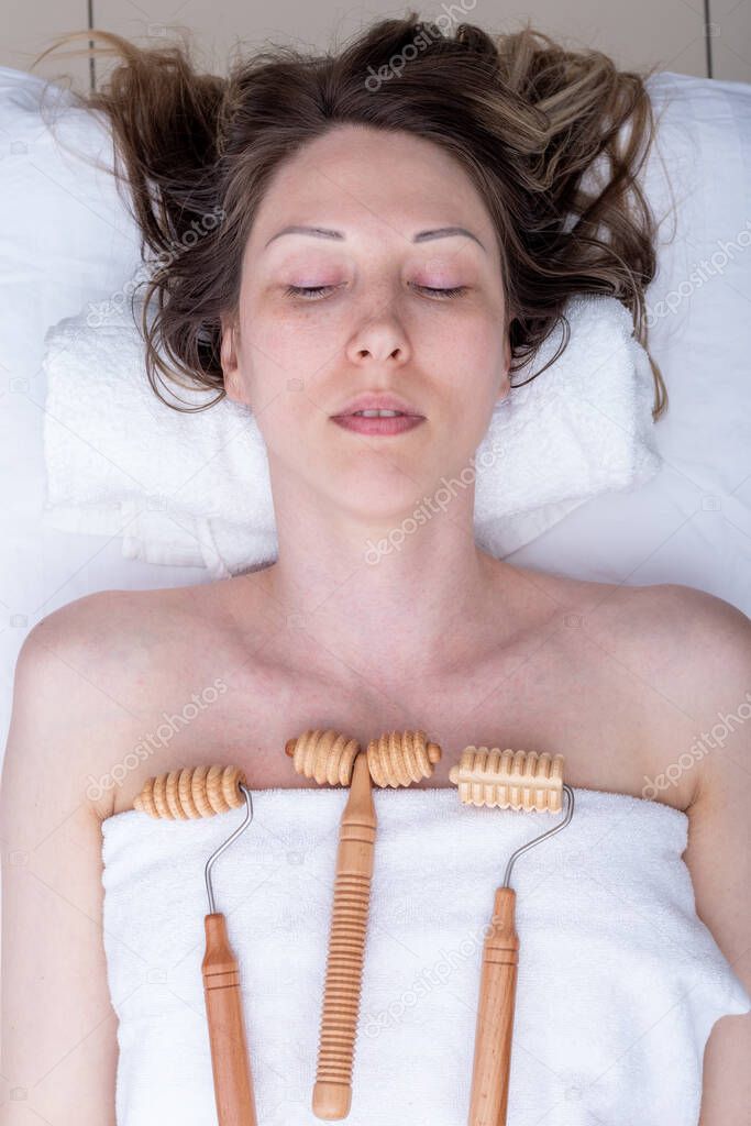 Madero therapy, anti-cellulite relaxing massage - wooden massage tools spiked rollers, smooth roller lying on a white towel on a woman, top view, close-up. Woman having spa massage at beauty salon.