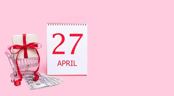 A gift box in a shopping trolley, dollars and a calendar with the date of 27 april on a pink background. — Stock Photo, Image