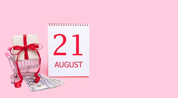 En presentask i en kundvagn, dollar och en kalender med datumet 21 augusti på en rosa bakgrund. — Stockfoto