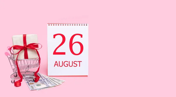 En presentask i en kundvagn, dollar och en kalender med datum 26 augusti på en rosa bakgrund. — Stockfoto