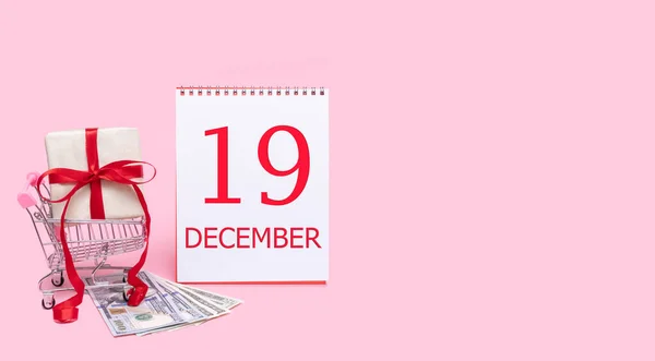 Presentförpackning i kundvagn, dollar och kalender med datum den 19 december på rosa bakgrund. — Stockfoto