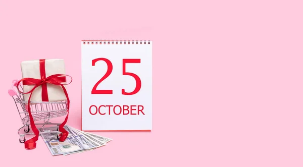 A gift box in a shopping trolley, dollars and a calendar with the date of 25 october on a pink background. — Stockfoto